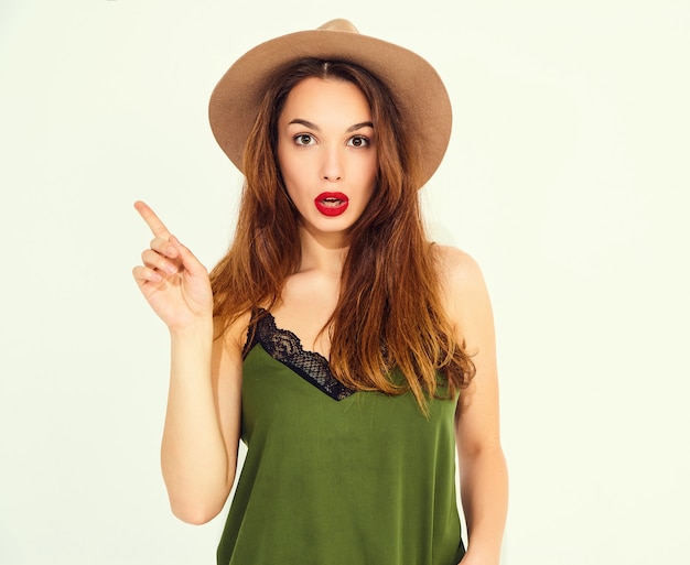 Młoda stylowa modelka w przypadkowych letnich zielonych ubraniach i brązowym kapeluszu z czerwonymi ustami wpadła na pomysł i podniosła palec. Odosobniony