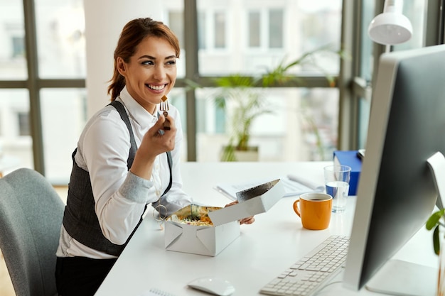 Młoda szczęśliwa bizneswoman korzysta z komputera podczas przerwy na lunch w biurze