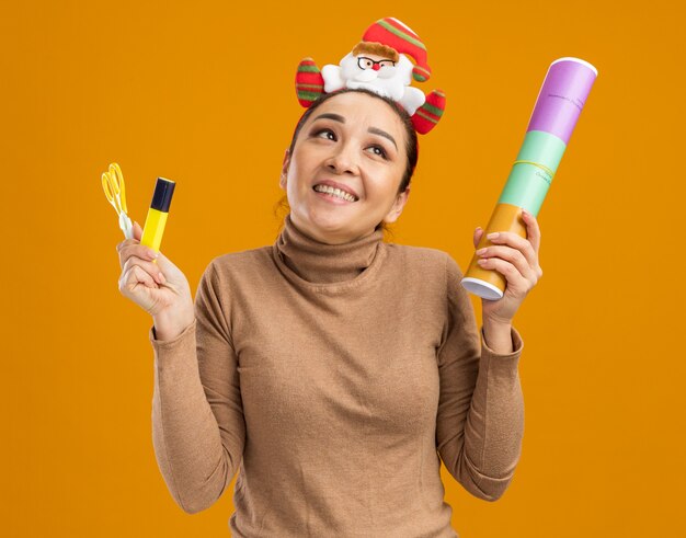 Młoda szczęśliwa dziewczyna w śmiesznej świątecznej obręczy na głowie trzymająca nożyczki petarda i klej