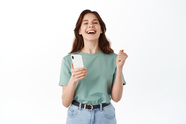 Młoda szczęśliwa kobieta mówi tak, wygrywając nagrodę lub codzienny cel w aplikacji mobilnej, trzymając smartfona i radując się, świętując zwycięstwo w aplikacji, stojąc na białym tle