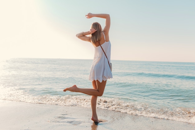 Młoda szczęśliwa kobieta tańczy odwracając się nad morzem na plaży w słoneczny letni styl mody w białej sukni wakacje