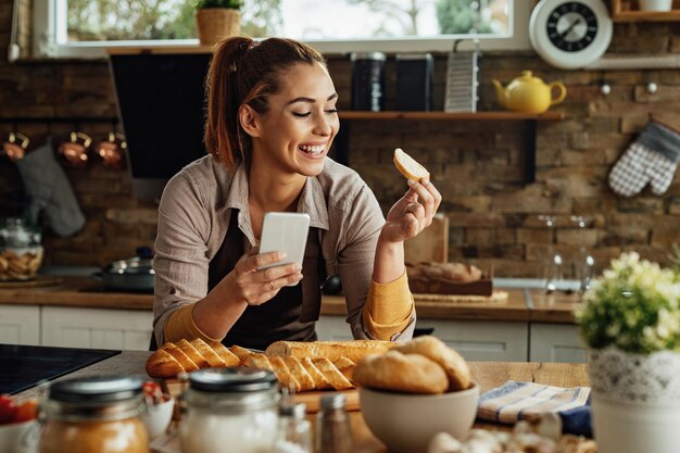 Młoda szczęśliwa kobieta trzymająca kromkę chleba podczas korzystania ze smartfona i przygotowywania jedzenia w kuchni