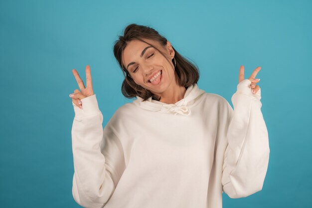 Młoda szczęśliwa kobieta w białej bluzie z kapturem radośnie pozuje na niebiesko w studio