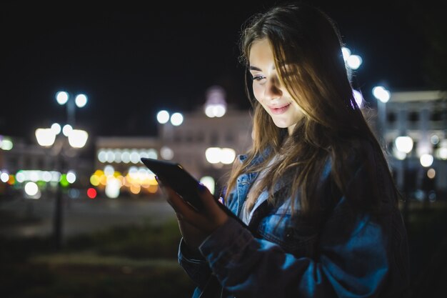 Młoda szczęśliwa kobieta za pomocą tabletu na zewnątrz przez rozmyte nocne światła miasta