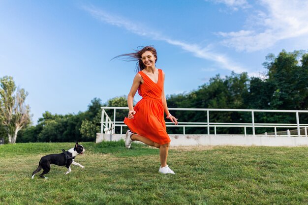 Młoda szczęśliwa uśmiechnięta kobieta w pomarańczowej sukience zabawy bawiąc się bieganiem z psem w parku, styl lato, wesoły nastrój