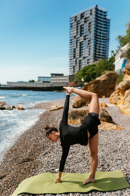 Młoda szczupła kobieta robi ćwiczenia na matę do jogi na zewnątrz przy kamienistej plaży nad morzem