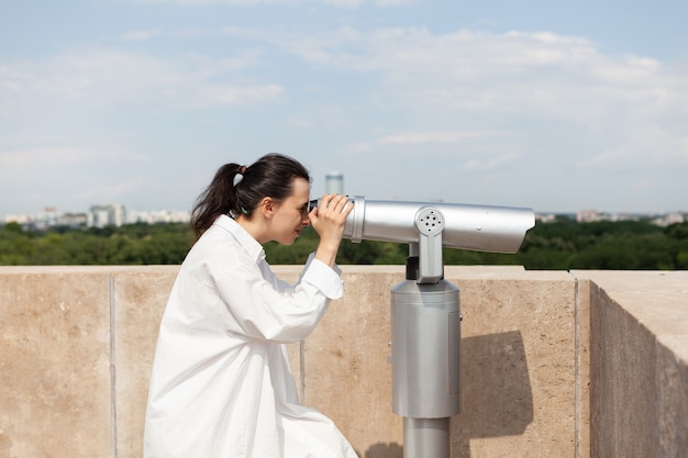 Bezpłatne zdjęcie młoda turystka stojąca na dachu budynku patrząca przez teleskop