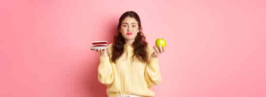 Bezpłatne zdjęcie młoda urocza kobieta wahająca się między pysznym kawałkiem ciasta deserem a zielonym zdrowym jabłkiem na diecie stojącej niezdecydowanie na różowym tle