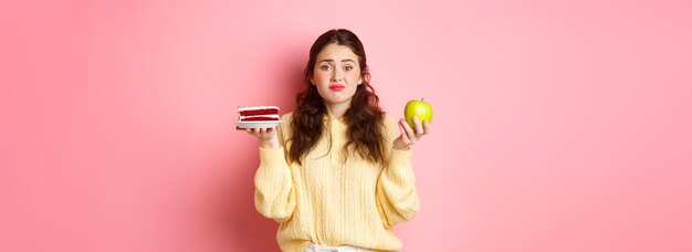 Młoda urocza kobieta wahająca się między pysznym kawałkiem ciasta deserem a zielonym zdrowym jabłkiem na diecie stojącej niezdecydowanie na różowym tle