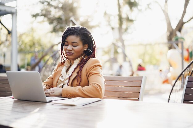 Młoda uśmiechnięta afroamerykanka siedzi przy stole w ulicznej kawiarni i korzysta z laptopa