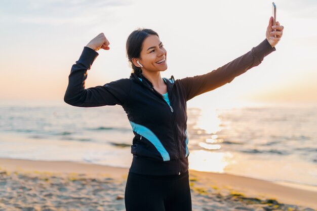 Młoda uśmiechnięta atrakcyjna szczupła kobieta robi ćwiczenia sportowe na plaży o poranku wschód słońca w strojach sportowych, zdrowy styl życia, słuchanie muzyki na słuchawkach, robienie zdjęć selfie na telefonie wyglądającym silnie