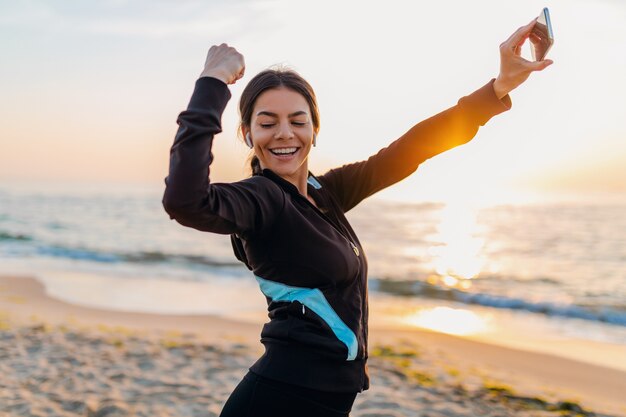 Młoda uśmiechnięta atrakcyjna szczupła kobieta robi ćwiczenia sportowe na plaży o poranku wschód słońca w strojach sportowych, zdrowy styl życia, słuchanie muzyki na słuchawkach, robienie zdjęć selfie na telefonie wyglądającym silnie