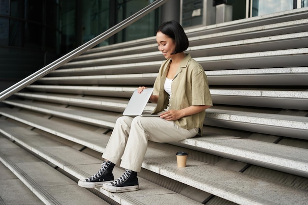 Bezpłatne zdjęcie młoda uśmiechnięta azjatka z laptopem siedzi na ulicy miasta z kawą młoda kobieta odrabia pracę domową dalej