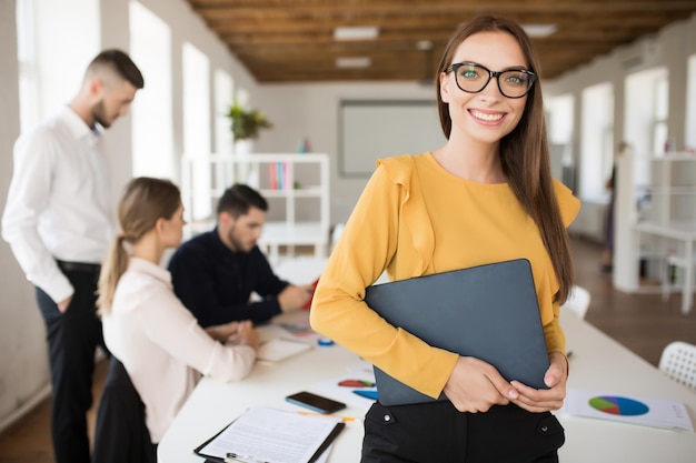 Bezpłatne zdjęcie młoda uśmiechnięta biznesowa kobieta w okularach szczęśliwie patrząca w kamerę, trzymając folder w rękach w biurze z kolegami w tle