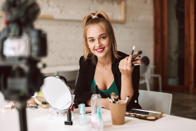 Młoda uśmiechnięta blogerka piękności szczęśliwie patrząca w kamerę trzymająca pędzle do makijażu w dłoni podczas nagrywania nowego wideo na vlog