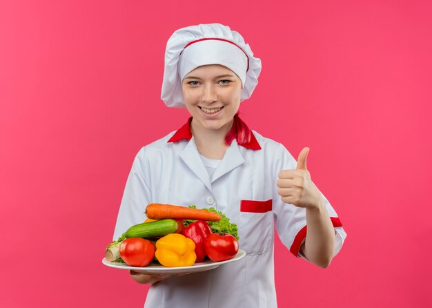 Młoda uśmiechnięta blondynka kobieta szef kuchni w mundurze szefa kuchni trzyma warzywa na talerzu i kciuki do góry na białym tle na różowej ścianie