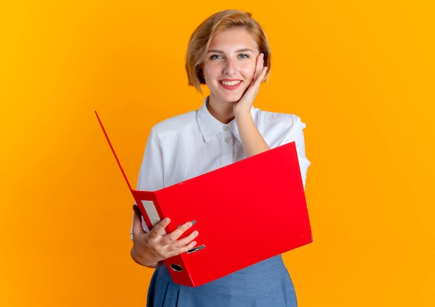 Młoda uśmiechnięta blondynka Rosjanka kładzie rękę na twarzy gospodarstwa folderu plików na białym tle na pomarańczowym tle z miejsca kopiowania