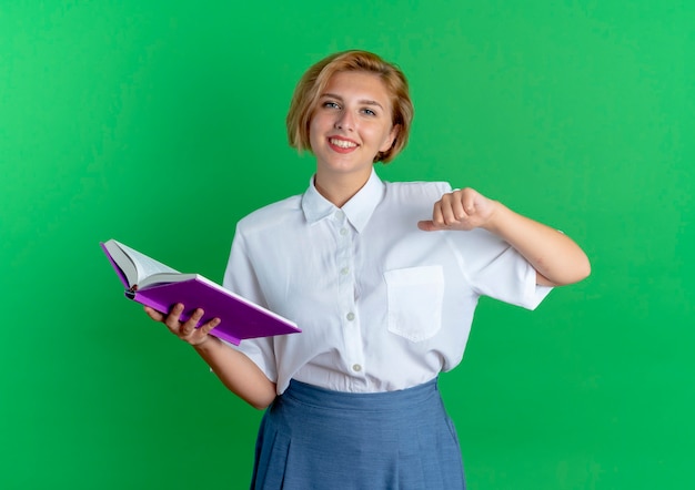 Bezpłatne zdjęcie młoda uśmiechnięta blondynka rosjanka trzyma książkę i wskazuje na siebie na białym tle na zielonym tle z miejsca na kopię