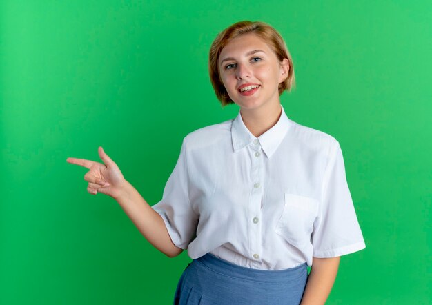 Bezpłatne zdjęcie młoda uśmiechnięta blondynka rosjanka wskazuje na bok patrząc na kamery na białym tle na zielonym tle z miejsca na kopię