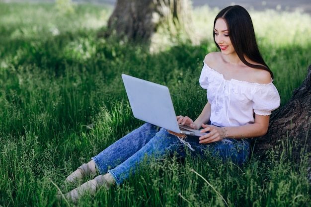 Młoda uśmiechnięta dziewczyna siedzi na trawie z laptopem na nogach