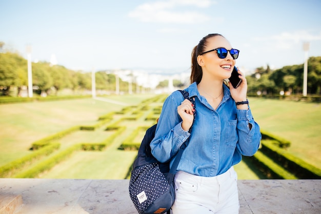 Młoda uśmiechnięta dziewczyna stoi w parku w okularach przeciwsłonecznych, odwracając wzrok i rozmawiając przez telefon