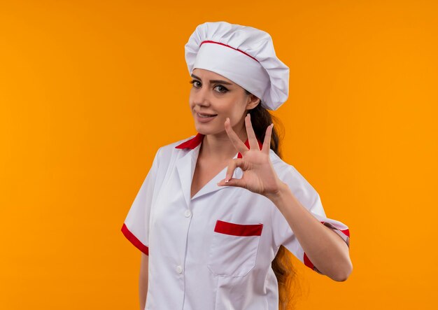 Młoda uśmiechnięta dziewczynka kaukaski kucharz w mundurze szefa kuchni gestykuluje ok ręka znak na białym tle na pomarańczowej ścianie z miejsca na kopię