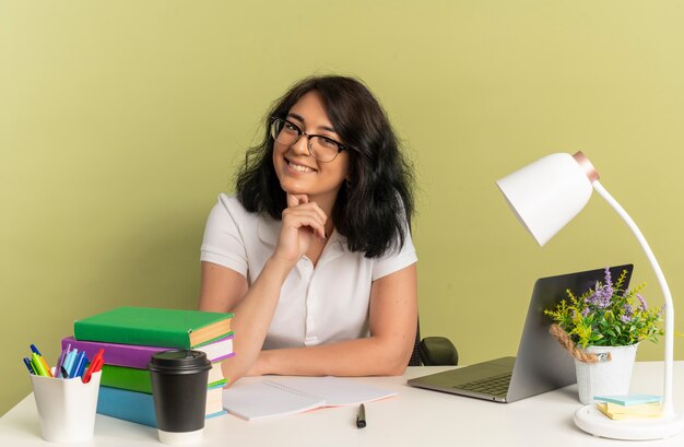 Młoda uśmiechnięta ładna kaukaska uczennica w okularach siedzi przy biurku z narzędziami szkolnymi kładzie dłoń na brodzie odizolowana na zielonej przestrzeni z miejscem na kopię