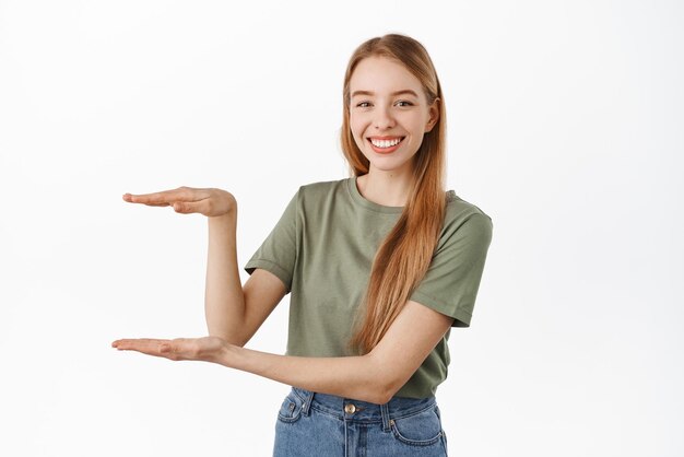 Młoda uśmiechnięta modelka pokazująca przedmiot trzymający coś pustego między dłońmi, wyglądająca na zadowoloną, polecając produkt stojący na białym tle