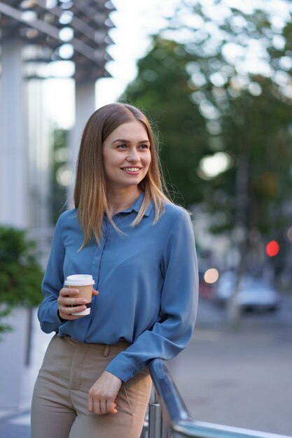 Młoda uśmiechnięta profesjonalna kobieta o przerwie na kawę podczas jej pełnego dnia pracy. Trzyma papierowy kubek na zewnątrz w pobliżu budynku biznesowego, jednocześnie relaksując się i delektując napojem.