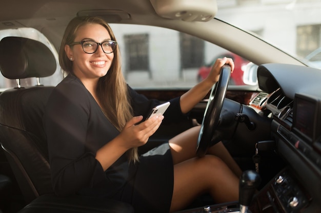 Młoda wesoła kobieta w czarnej sukience i okularach szczęśliwie patrząc w aparacie z telefonem komórkowym w ręku siedząc za kierownicą w samochodzie