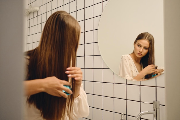 Młoda wspaniała kobieta w białej sukni w zamyśleniu szczotkuje włosy w pobliżu lustra w łazience