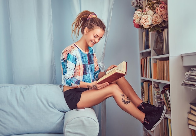 Młoda wytatuowana dziewczyna czyta książkę siedząc na kanapie w domu.