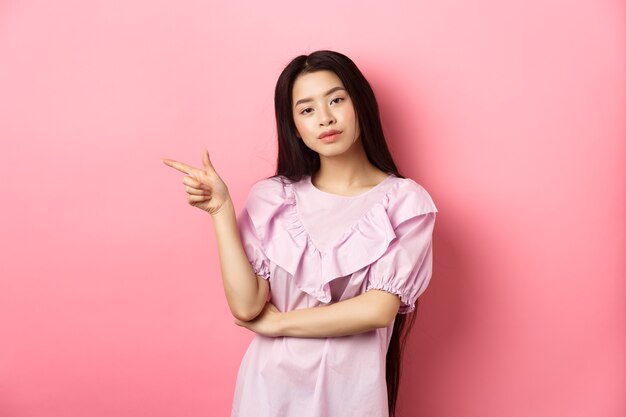 Młoda zadowolona Azjatka wygląda fajnie i wskazuje palcem w lewo na logo produktu reklamowego na różowym roma...