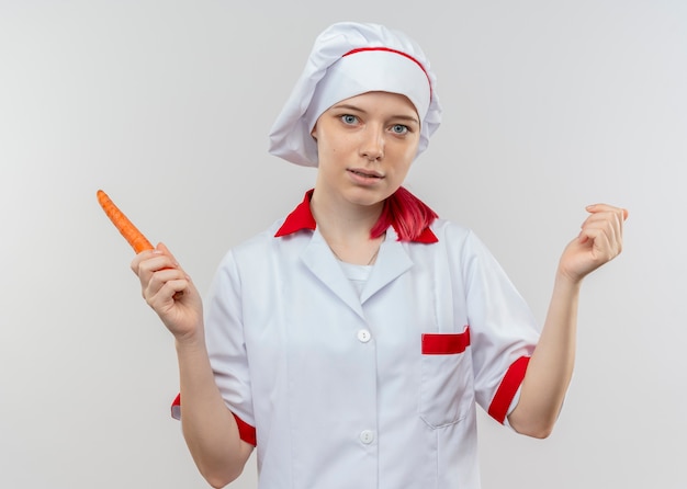 Młoda zadowolona blondynka szef kuchni w mundurze szefa kuchni trzyma marchewkę i wygląda na białym tle na białej ścianie