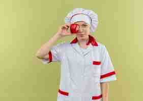 Bezpłatne zdjęcie młoda zadowolona blondynka szef kuchni w mundurze szefa kuchni zamyka oko z czerwonym pieprzem na białym tle na zielonej ścianie