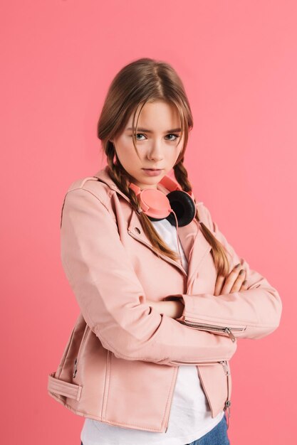 Młoda zdenerwowana dziewczyna z dwoma warkoczami w skórzanej kurtce ze słuchawkami na szyi, trzymając się za rękę, smutno patrząc w kamerę na różowym tle