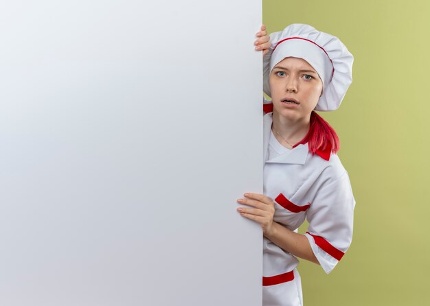 Młoda zdezorientowana blondynka kobieta szef kuchni w mundurze szefa kuchni stoi i patrzy za białą ścianę na białym tle na zielonej ścianie