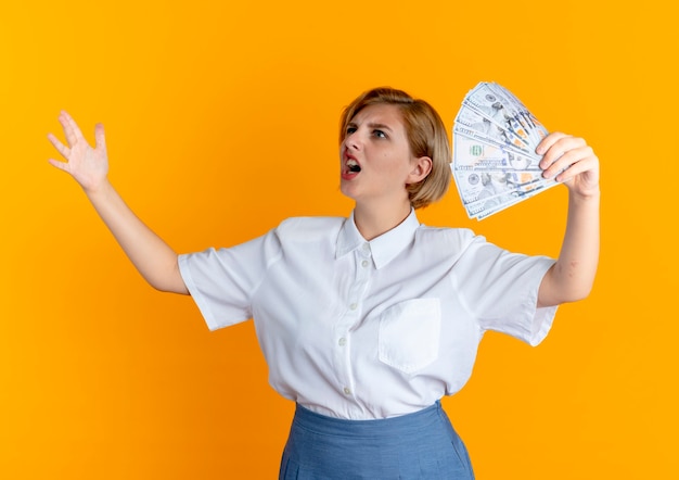 Młoda zirytowana blondynka Rosjanka trzyma pieniądze, patrząc na bok z podniesioną ręką na białym tle na pomarańczowym tle z miejsca na kopię