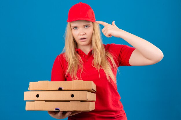 Młoda znudzona kobieta dostawy na sobie czerwoną koszulkę polo i czapkę stojącą z pudełkami po pizzy, strzelając do siebie, robiąc palec z pistoletu podpisując na odosobnionej niebieskiej ścianie