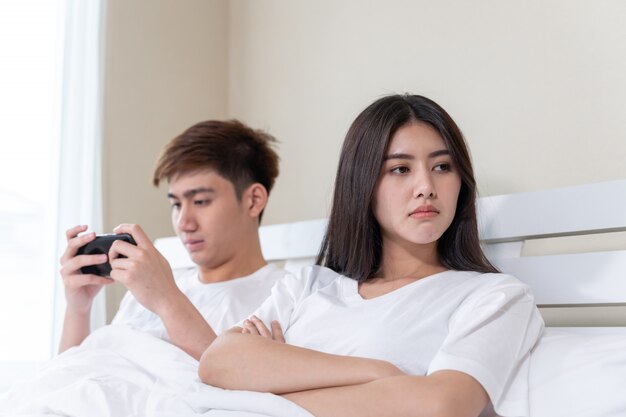 młoda żona siedzi na łóżku z uczuciem zdenerwowania, jej mąż używa smartfona w czasie drzemki