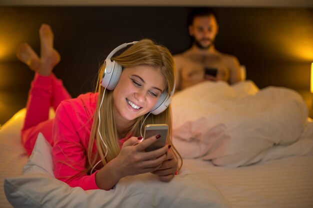 Młode szczęśliwe kobiety na białym łóżku przy użyciu telefonu komórkowego ze słuchawkami, chłopak na tle. Pojęcie o technologii i ludziach