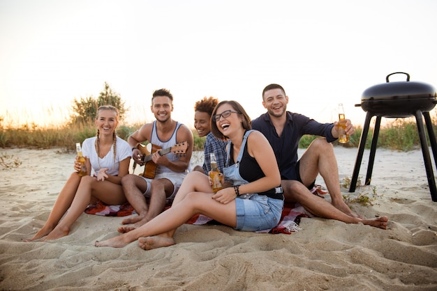 Młode towarzystwo przyjaciół radujących się, odpoczywających na plaży podczas wschodu słońca