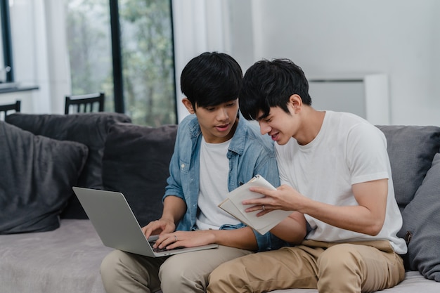 Młodej Azjatyckiej Homoseksualnej pary pracujący laptop przy nowożytnym domem. Asia LGBTQ + mężczyźni chętnie relaksują się przy użyciu komputera i analizują wspólnie swoje finanse w Internecie, leżąc na kanapie w salonie w domu.