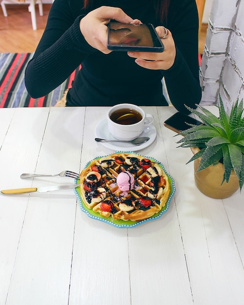 Bezpłatne zdjęcie młodej dziewczyny siedzący caffe je gofra śniadaniowego z czekoladowym sosem, plasterkami banana i truskawkami na zielonym ceramicznym talerzu i fotografował jej śniadanie