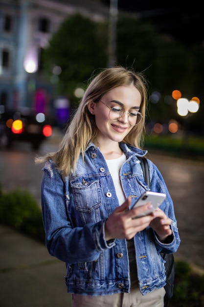 Młodej kobiety plenerowa noc używać mądrze telefon twarz iluminującą ekranem świetlnym. Internet, sieć społecznościowa, koncepcja technologii