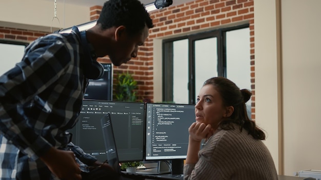 Bezpłatne zdjęcie młodszy programista siedzący na biurku, pokazujący laptop z kodem źródłowym starszemu programiście z prośbą o opinię. algorytm pisania programisty przerwany przez kolegę chcącego pomóc w naprawie błędów.