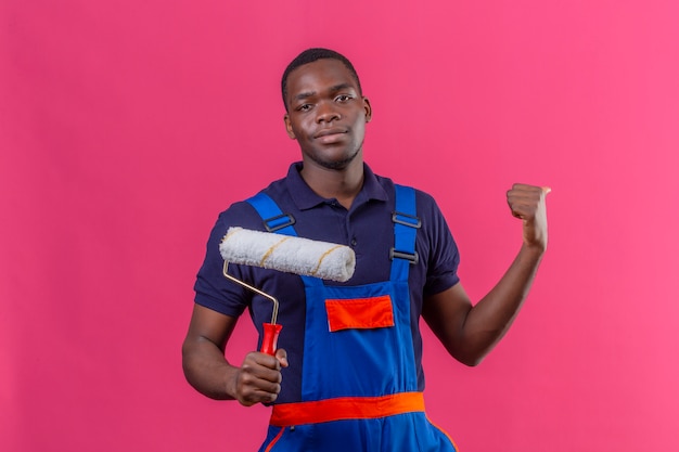 Młody African American budowniczy mężczyzna ubrany w mundur budowlany, trzymając wałek do malowania wskazując kciukiem z powrotem z kciukiem, patrząc pewnie stojąc na różowo