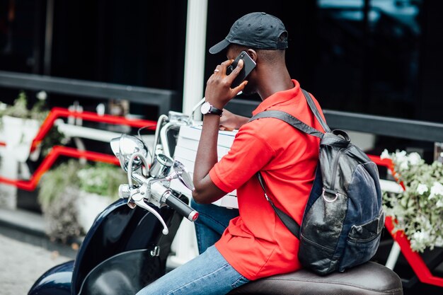 Młody Afrykanin przyjmuje zamówienie przez telefon i pisze w motocyklu trzymając pudełka z pizzą