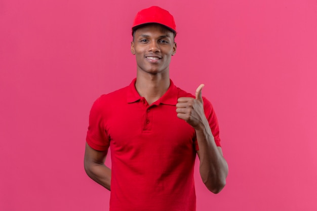 Młody amerykanin afrykańskiego pochodzenia doręczeniowy mężczyzna jest ubranym czerwoną polo koszula i nakrętkę z uśmiechem na twarzy pokazuje aprobaty nad odosobnionym menchiami