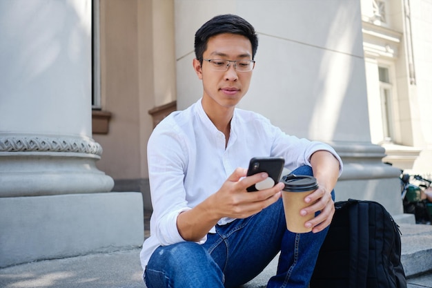 Młody Atrakcyjny Azjatycki Student Płci Męskiej W Okularach Z Kawą, Aby Ostrożnie Korzystać Z Telefonu Komórkowego W Pobliżu Uniwersytetu